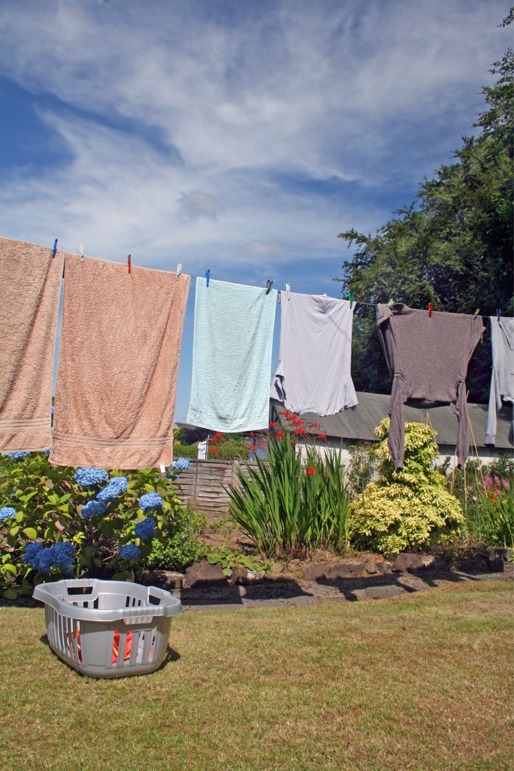 Laundry hanging on a line in a sunny garden, helping to lower energy costs, surrounded by vibrant flowers and a basket of clothes on the grass.