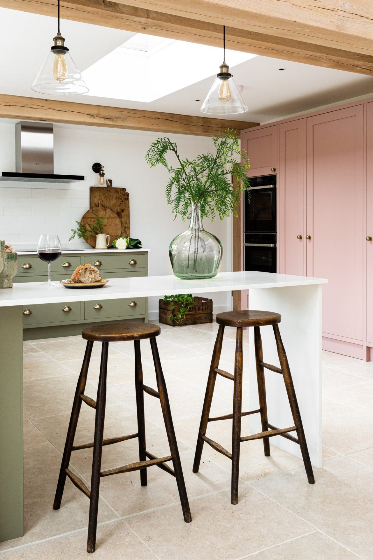 A stylish small kitchen with pink cabinetry, a white island with two wooden stools, hanging pendant lights, and a large glass vase with greenery.