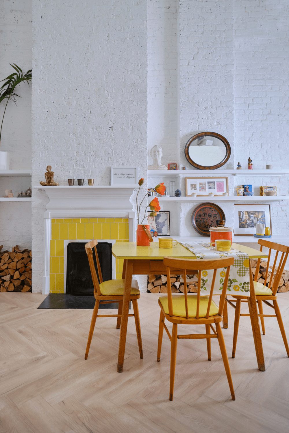 A cozy dining area with a yellow table and chairs, white brick walls, innovative flooring ideas for small spaces, a fireplace with stacked firewood, and assorted decor on the mantelpiece.