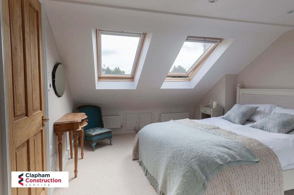 A bright, well-lit attic bedroom with skylights, featuring a neatly made bed and simple furnishings, accessed by stairs designed for a loft conversion.