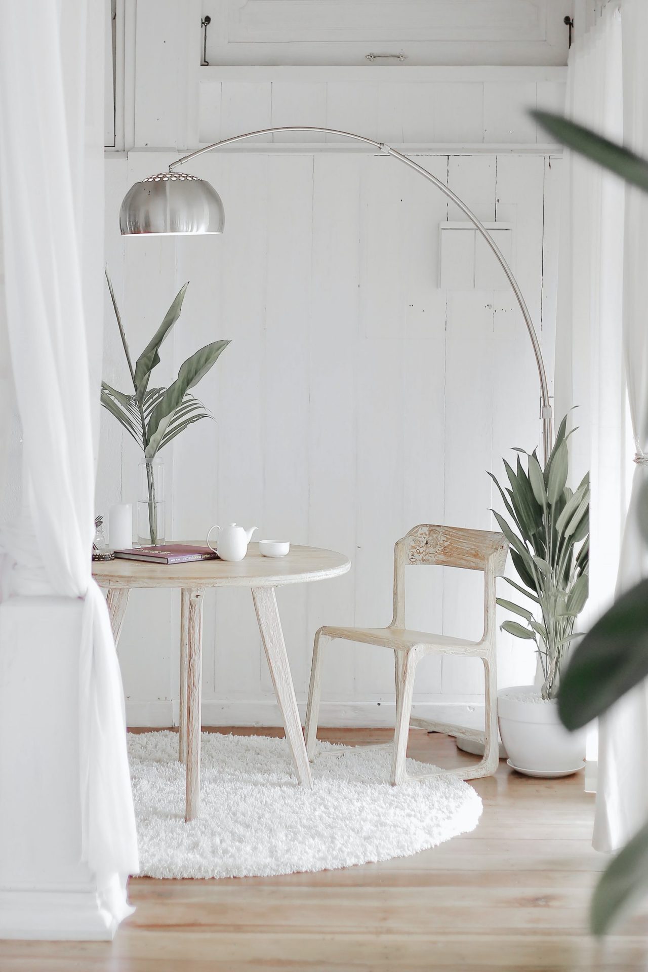 A minimalist living room in Japandi style with a white color scheme, featuring a round table, a single chair, a floor lamp, and some green potted plants.