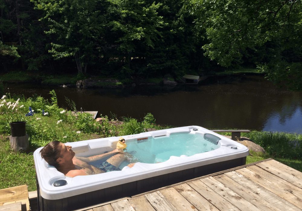 man lying in small hot tub that looks like a bath on the side of a lake