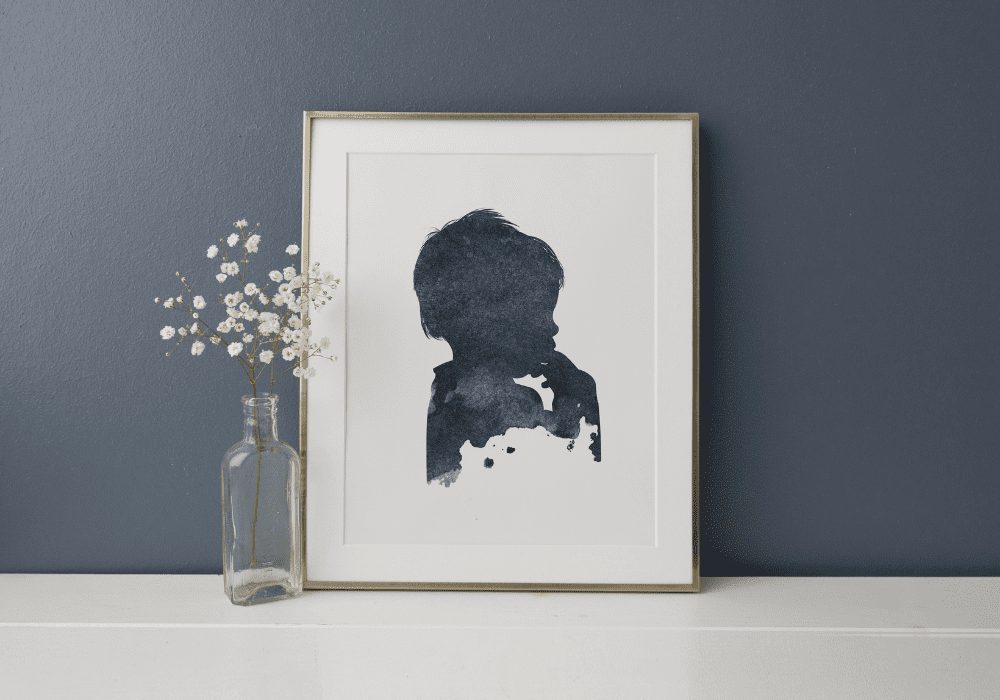 A silhouette painting of a child's profile in a burnt peach frame, placed on a shelf next to a glass vase with white flowers against a dark gray wall.