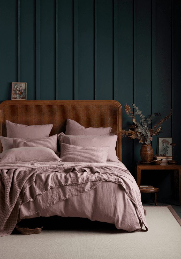 A neatly made bed with mauve bedding against a dark blue paneled wall in a dimly lit room, reflecting a budget bedroom design with minimal decor.