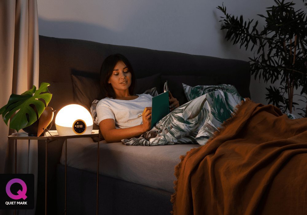 A woman reading a book on how to wake up refreshed, on a couch in a dimly lit room with a glowing table lamp displaying 9:29.