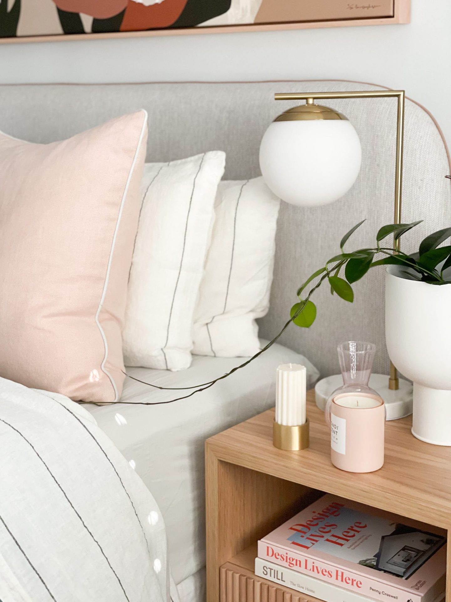A cozy bedroom reading nook with a soft pink pillow, white bedding, a bedside table featuring a sphere lamp, plants, and candles, accompanied by a bookshelf filled with books and decorative items to