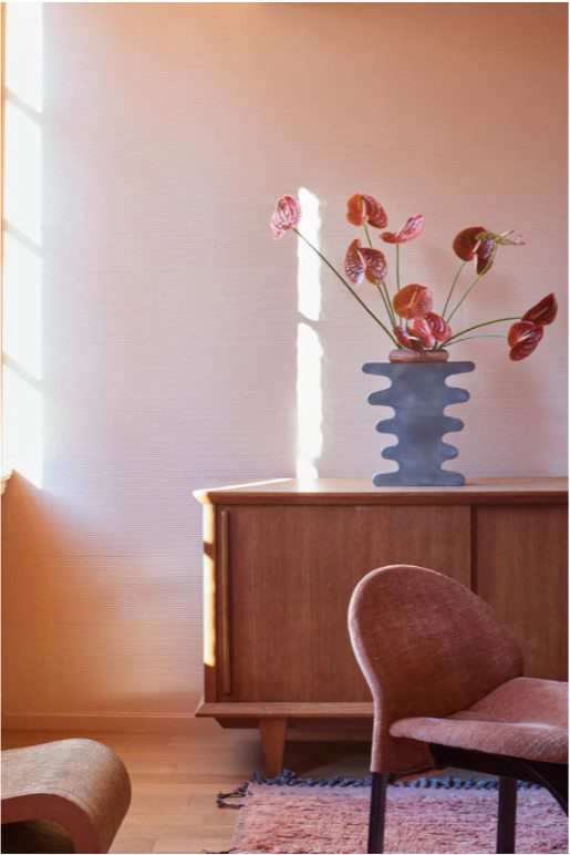 Farrow and ball paint terracotta on the walls behind a teak sideboard wtih a large vase of red flowers, pink rug on the floor and peachy coloured chair