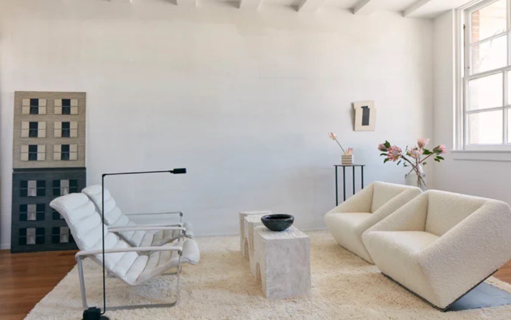 A setting for Farrow and ball paint, with pale white walls, modern white chairs around an off white rug, industrial window and no blinds
