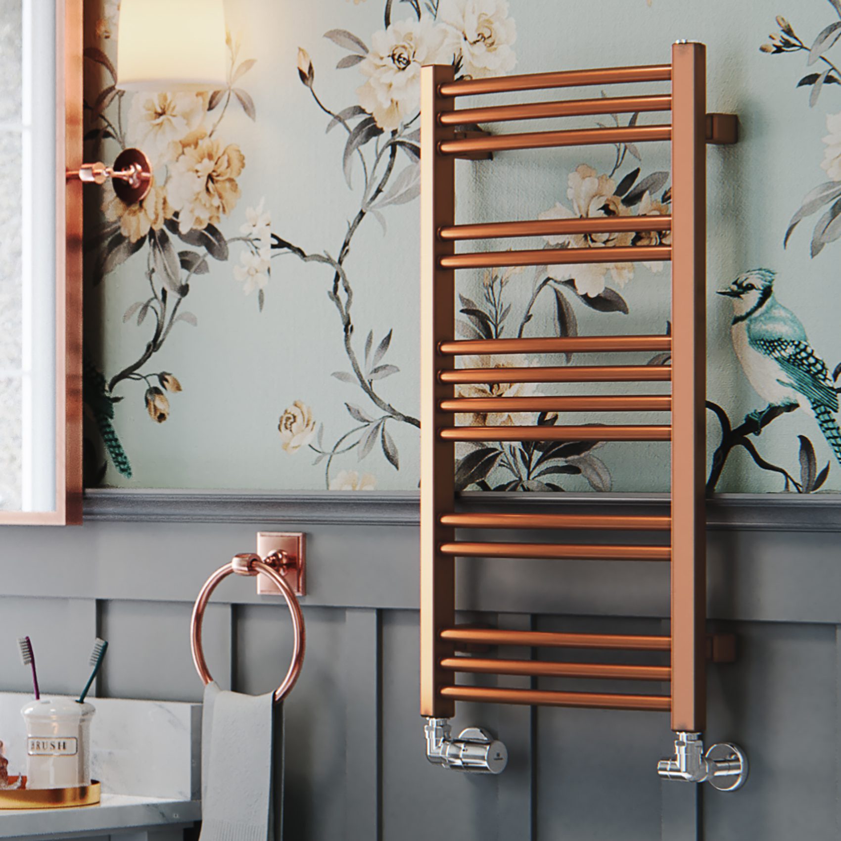 A copper towel radiator mounted on a wall with floral wallpaper, next to a mirror with a copper frame, and bathroom amenities on a shelf helps cut heating costs.