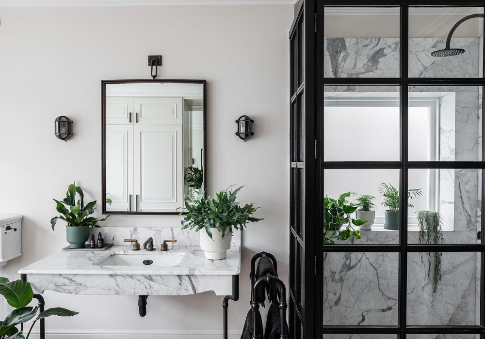 Elegant bathroom renovation featuring a marble vanity, wall-mounted mirror, black frame accents, and decorative plants.