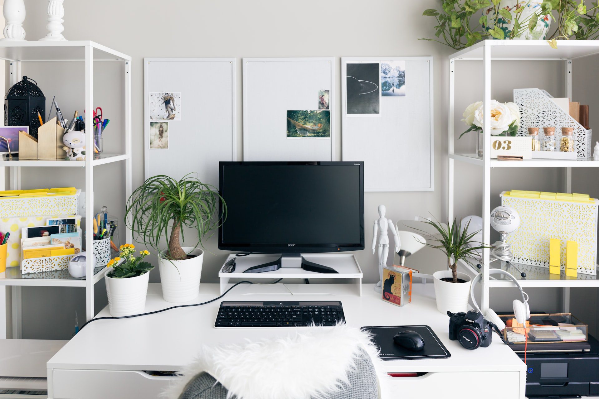 A tidy home office equipped for a budding online business with a desktop computer on a white desk, surrounded by shelves filled with books, boxes, plants, and decorative items.