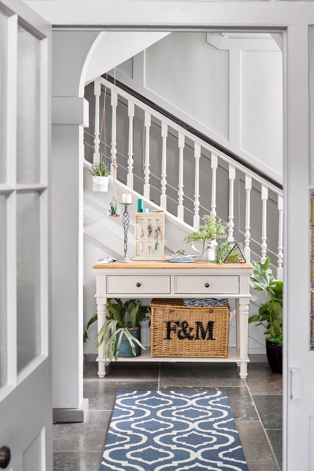 A hallway featuring a white staircase with a wooden console table adorned with plants and decor, a woven basket, and a blue patterned rug in a luxury home.