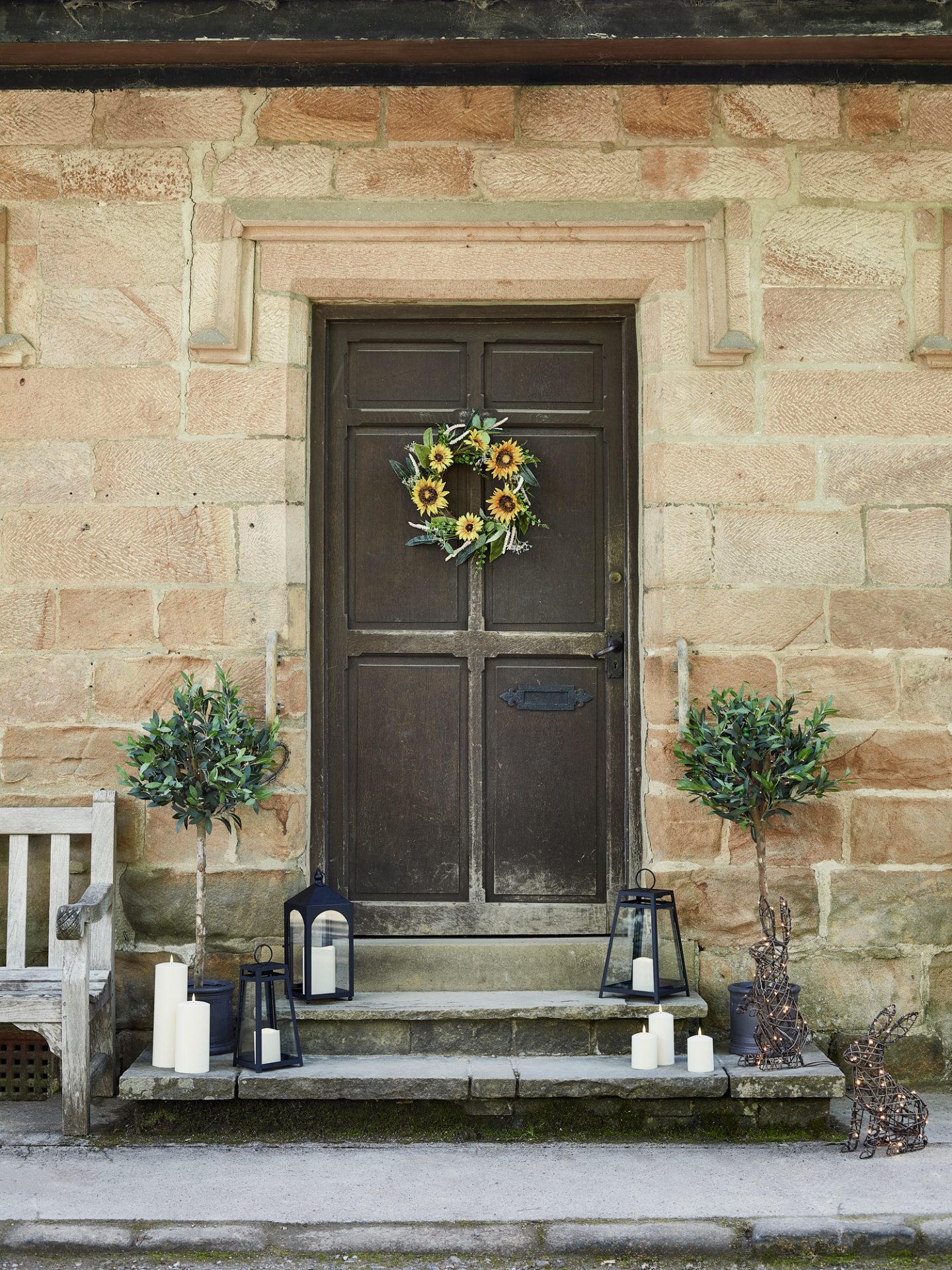 An old wooden front door adorned with a spectacular spring wreath, flanked by two potted plants and decorated with lanterns on the steps.