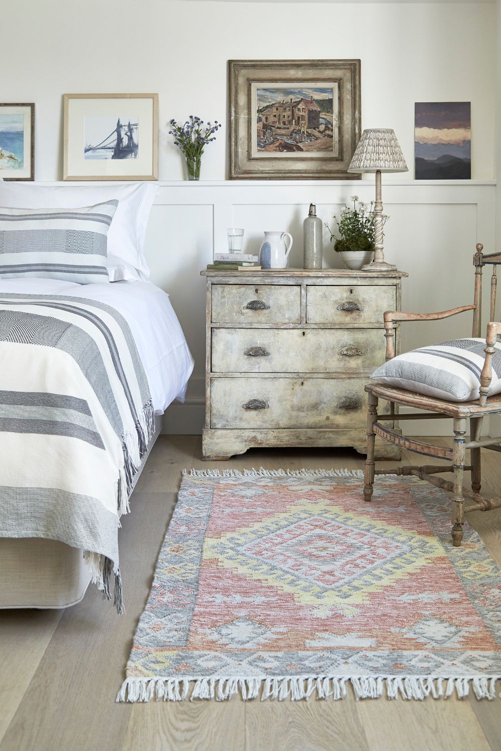 A cozy bedroom featuring a neatly made bed with striped bedding, a rustic wooden dresser, a small chair, and a colorful rug on a wooden floor, appreciated by elderly parents. Artwork hangs above the