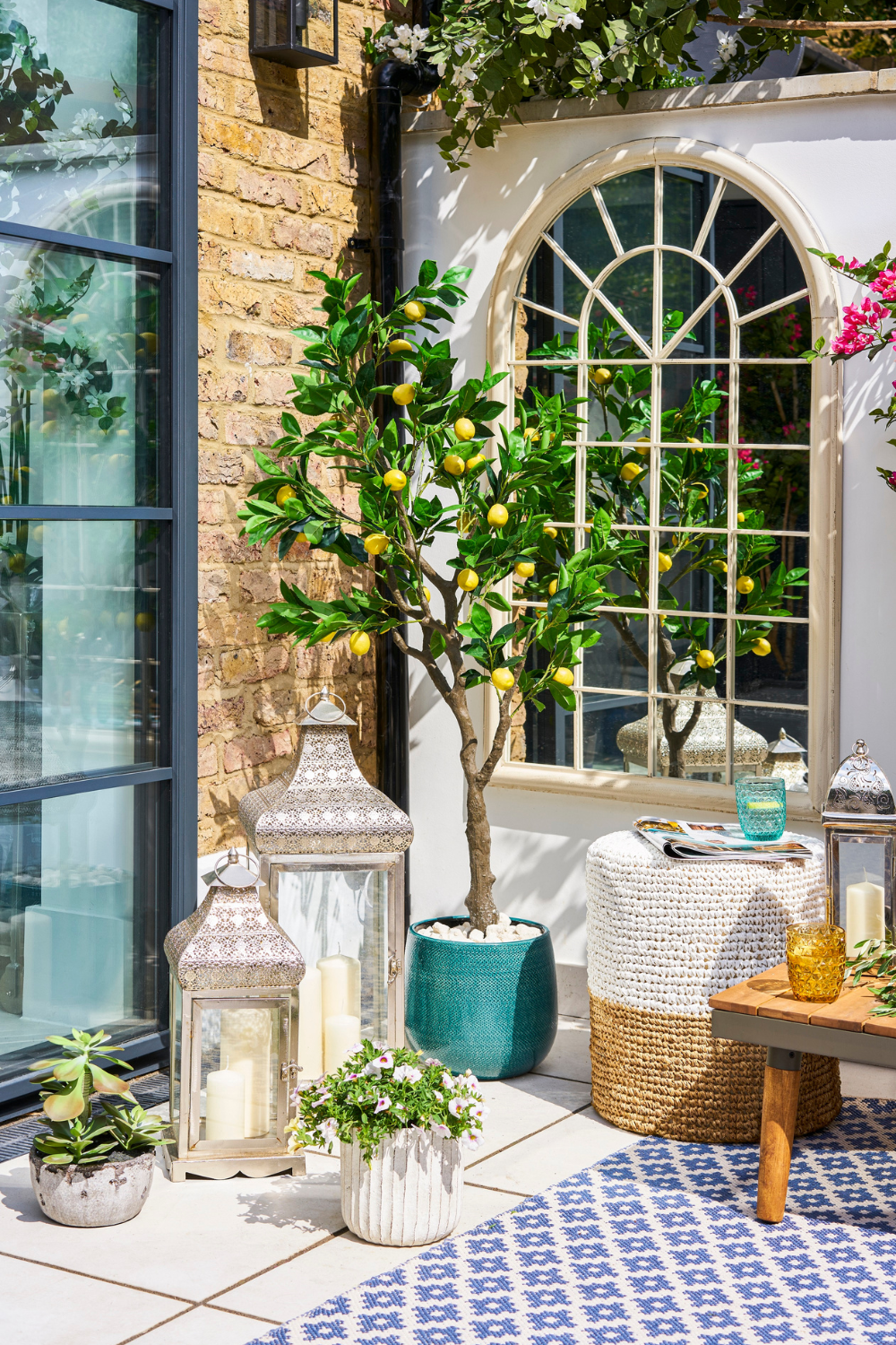 Sunny patio with a lemon tree, various lanterns, a woven ottoman, and a table set with glasses, surrounded by flowering plants and a patterned rug in a small garden.