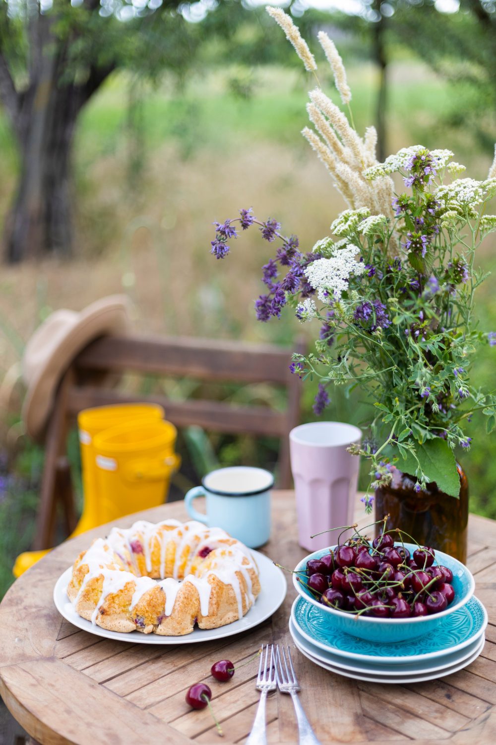 A rustic outdoor setting with a wooden table displaying a plate of scones, a bowl of cherries, a mug of coffee, and a colorful bouquet in a vase, perfect for a summer garden