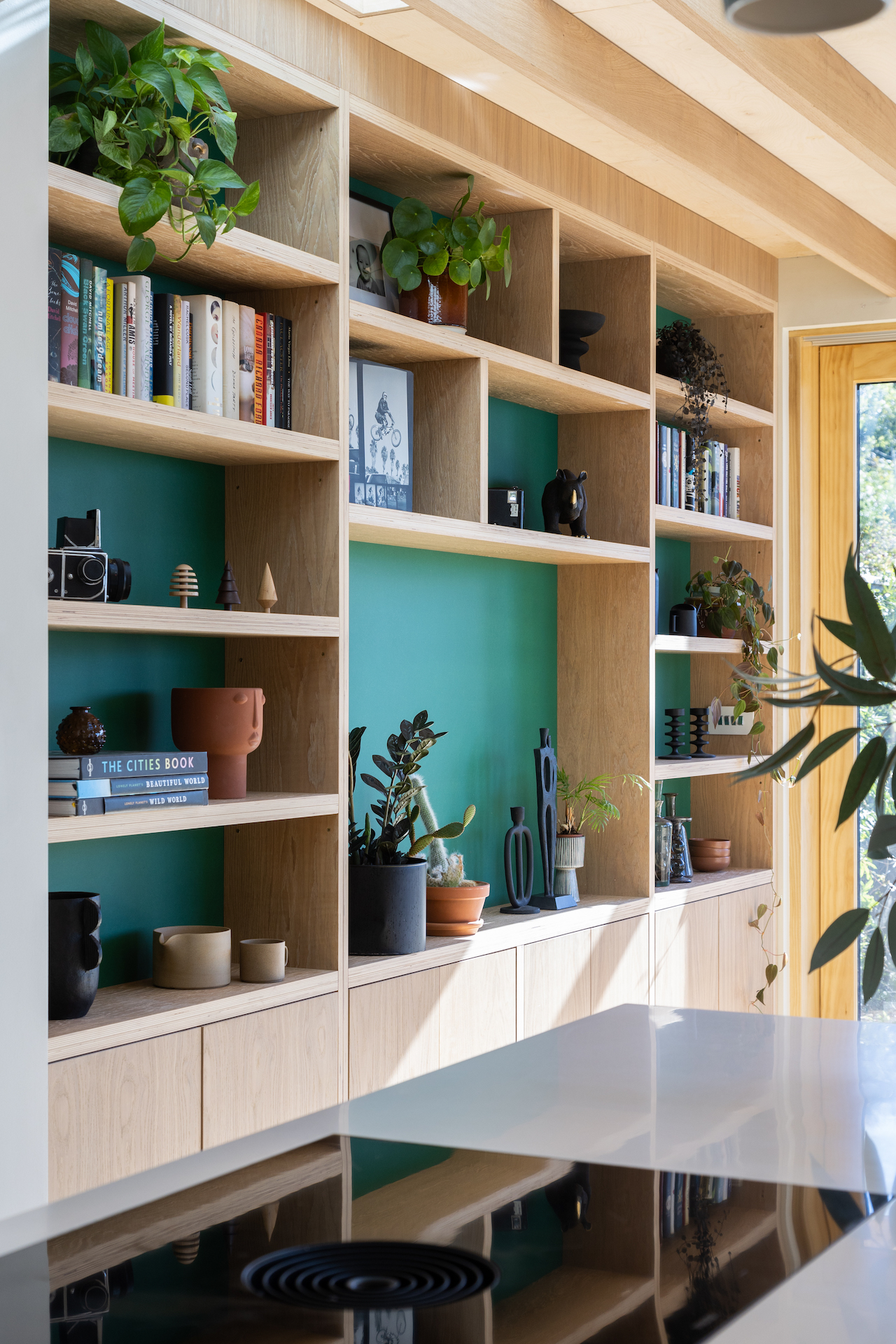 A bright, modern kitchen with a large built-in shelving unit painted teal, filled with books, plants, and decorative items, reflecting in a glossy countertop—resulting from a stress-free renovation