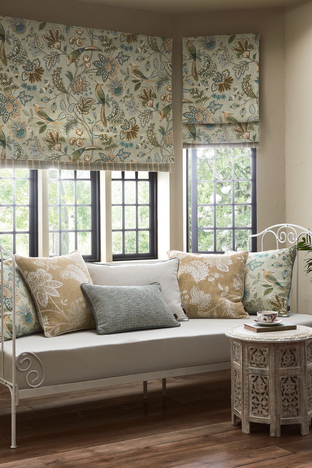 A cozy living room window seat with patterned cushions and matching floral roman blinds, beside a white metal table with a teapot and cup.