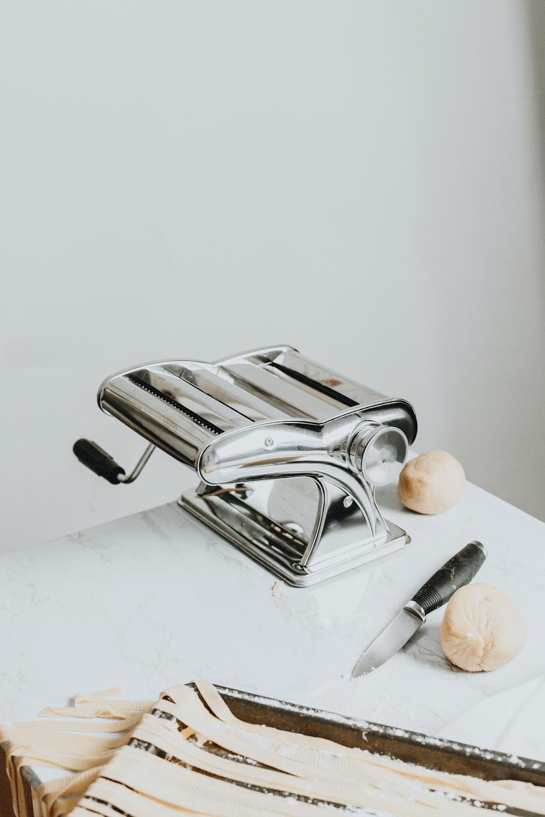 A pasta machine maker with a handle and clamp on a white table alongside a knife and two balls of dough, with fresh pasta strips partially visible in the foreground.