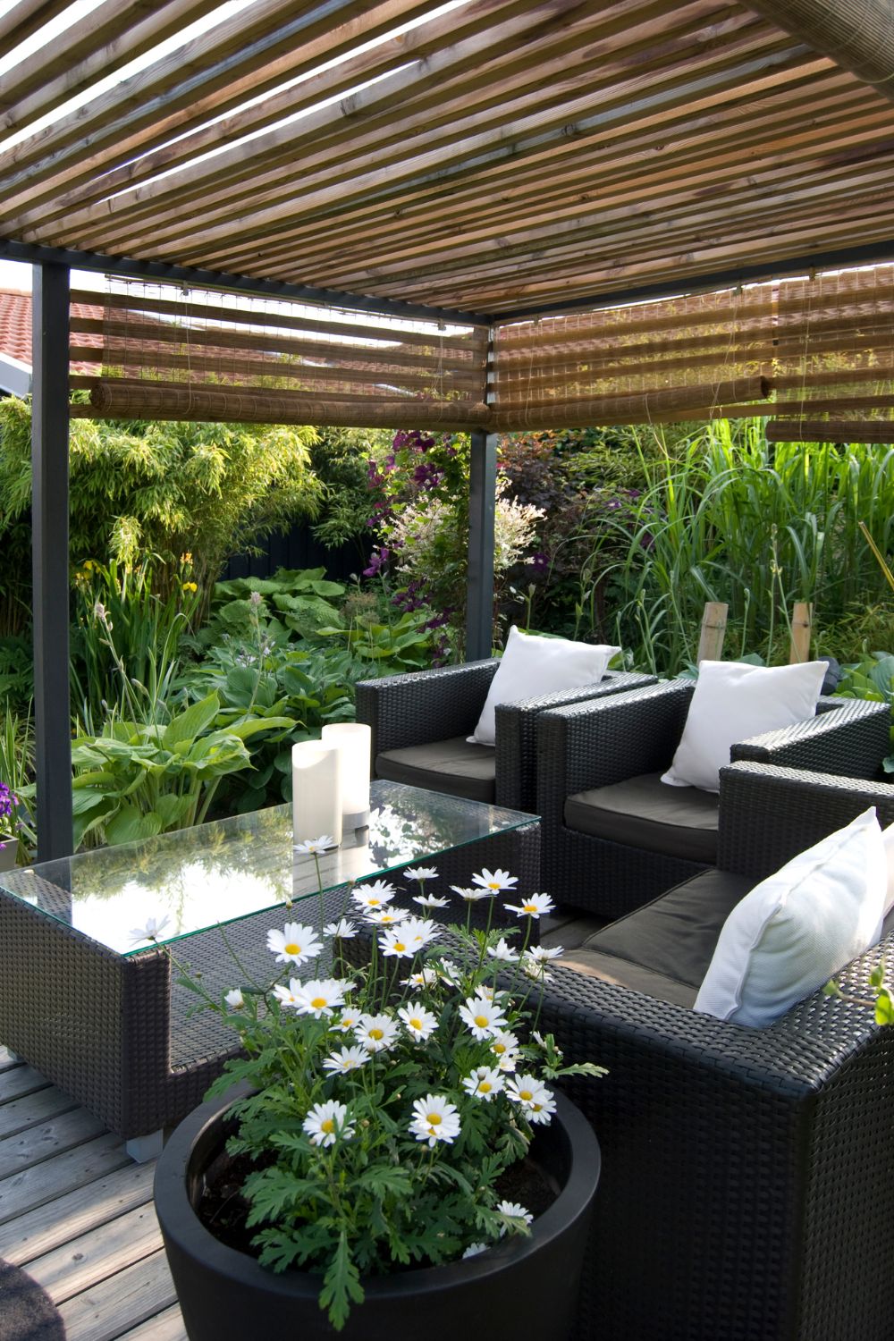 Outdoor patio with a pergola, featuring wicker furniture, a glass table, and decorative plants, surrounded by lush greenery freshly enhanced by a garden renovation.