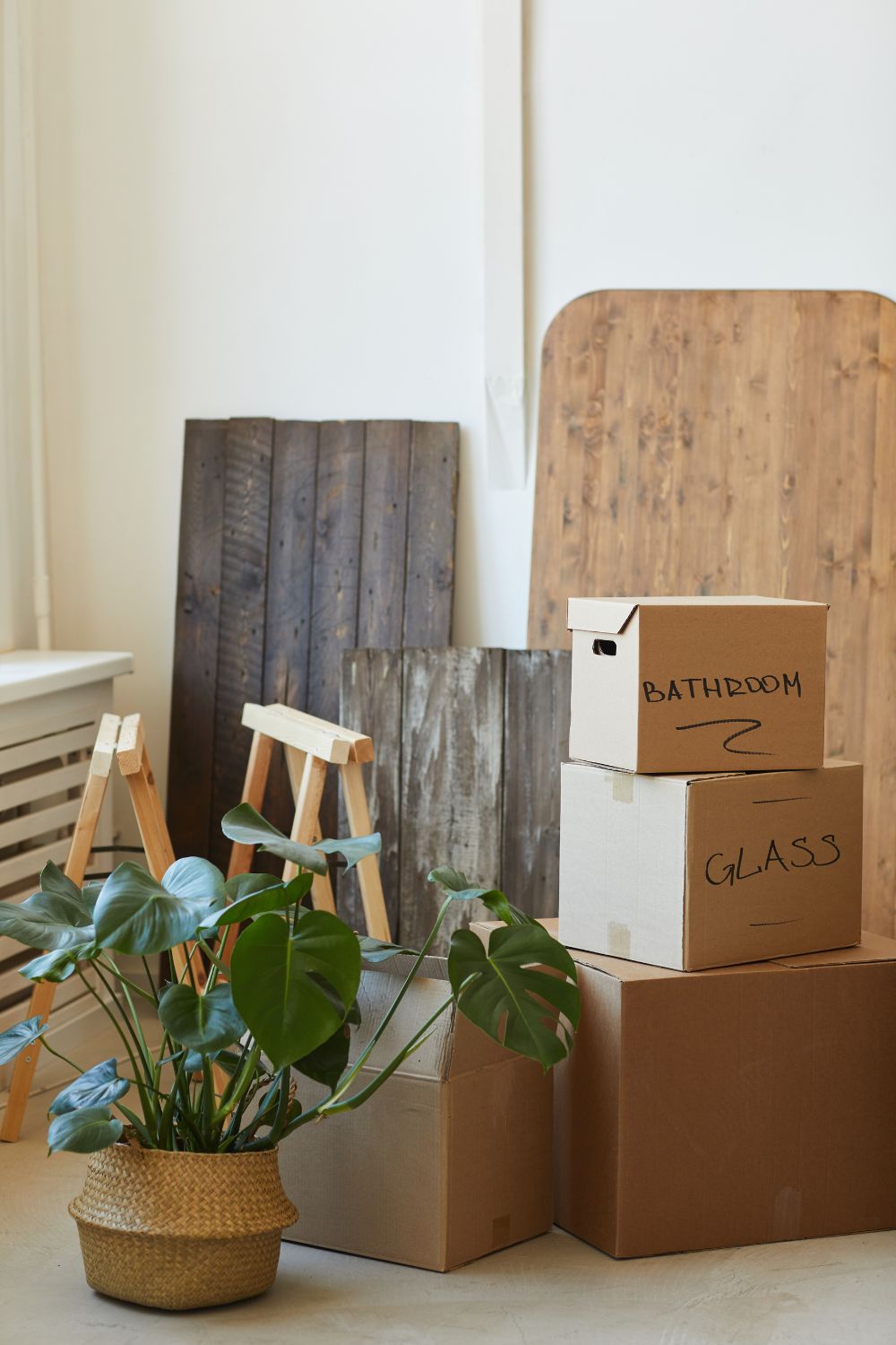 Stack of labeled moving boxes with "bathroom" and "glass" beside a potted plant in a room with wooden boards and easels, offering practical moving tips.