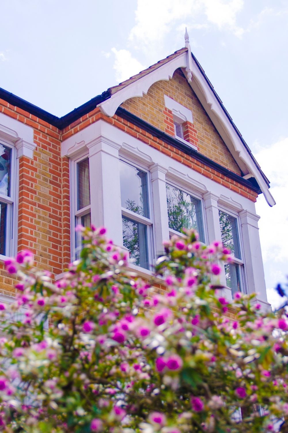 Victorian house with a gabled roof and red brickwork visible behind colorful blooming bushes under a cloudy sky, perfect for your next real estate investment.
