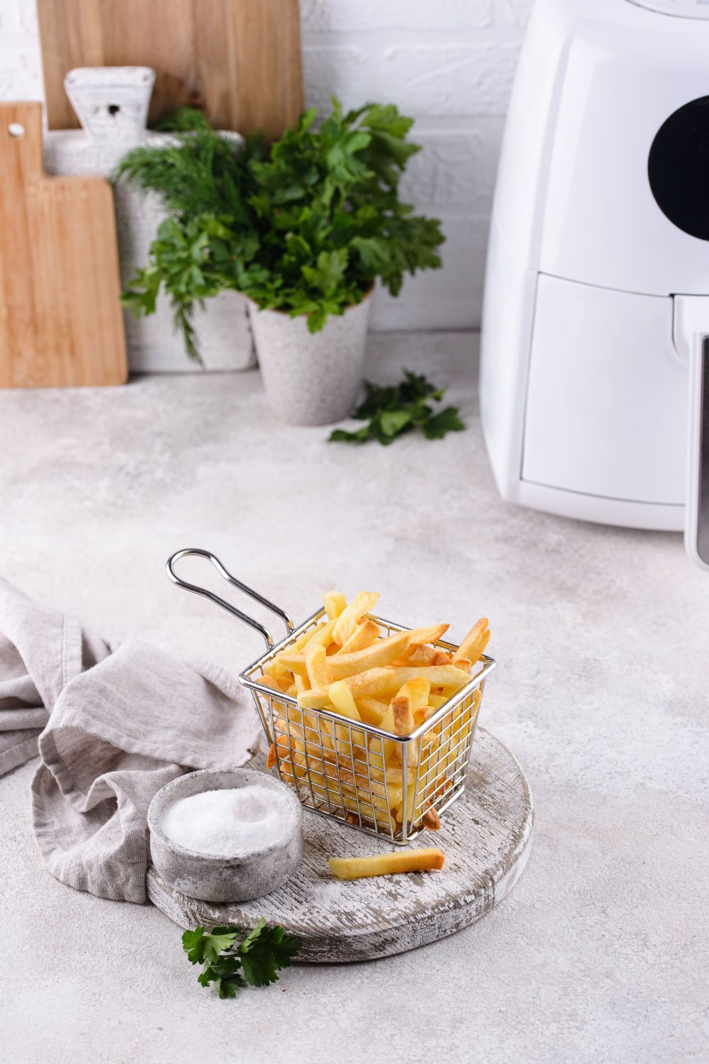 A small wire basket filled with crispy french fries, accompanied by a salt cellar, sits on a kitchen counter with an air fryer, one of the latest kitchen must-haves, in the background.