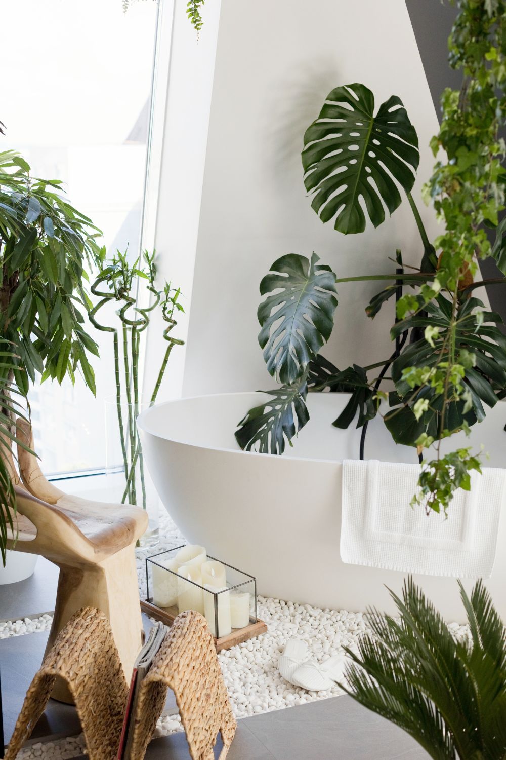Boho bathroom decor with white bath and lots of green plants, a white bath mat, and wicker magazine rack.