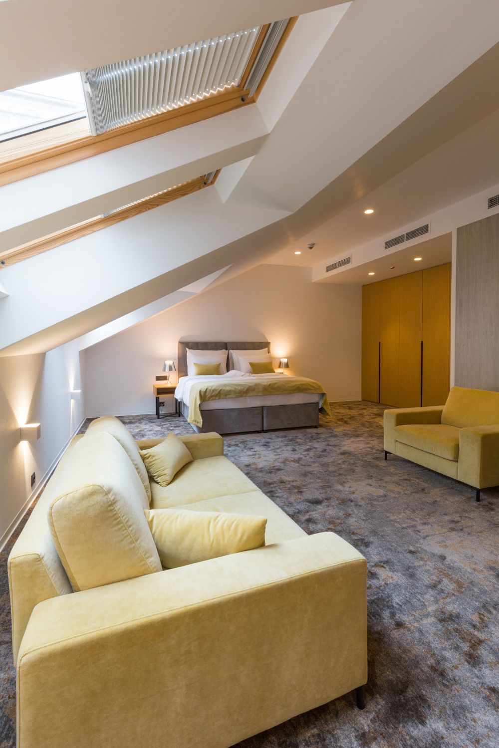 Modern attic bedroom with plush yellow sofas, a large bed, roof windows, wooden accents, and soft lighting.