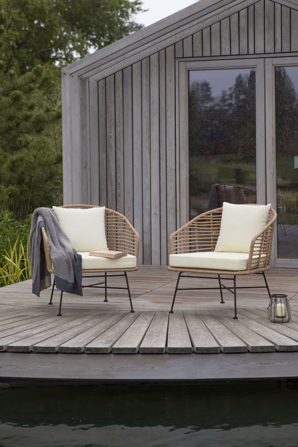 Two modern wicker chairs with white cushions on a freshly restained wooden deck by a serene lake, with a gray blanket on one chair and a small lantern beside them.