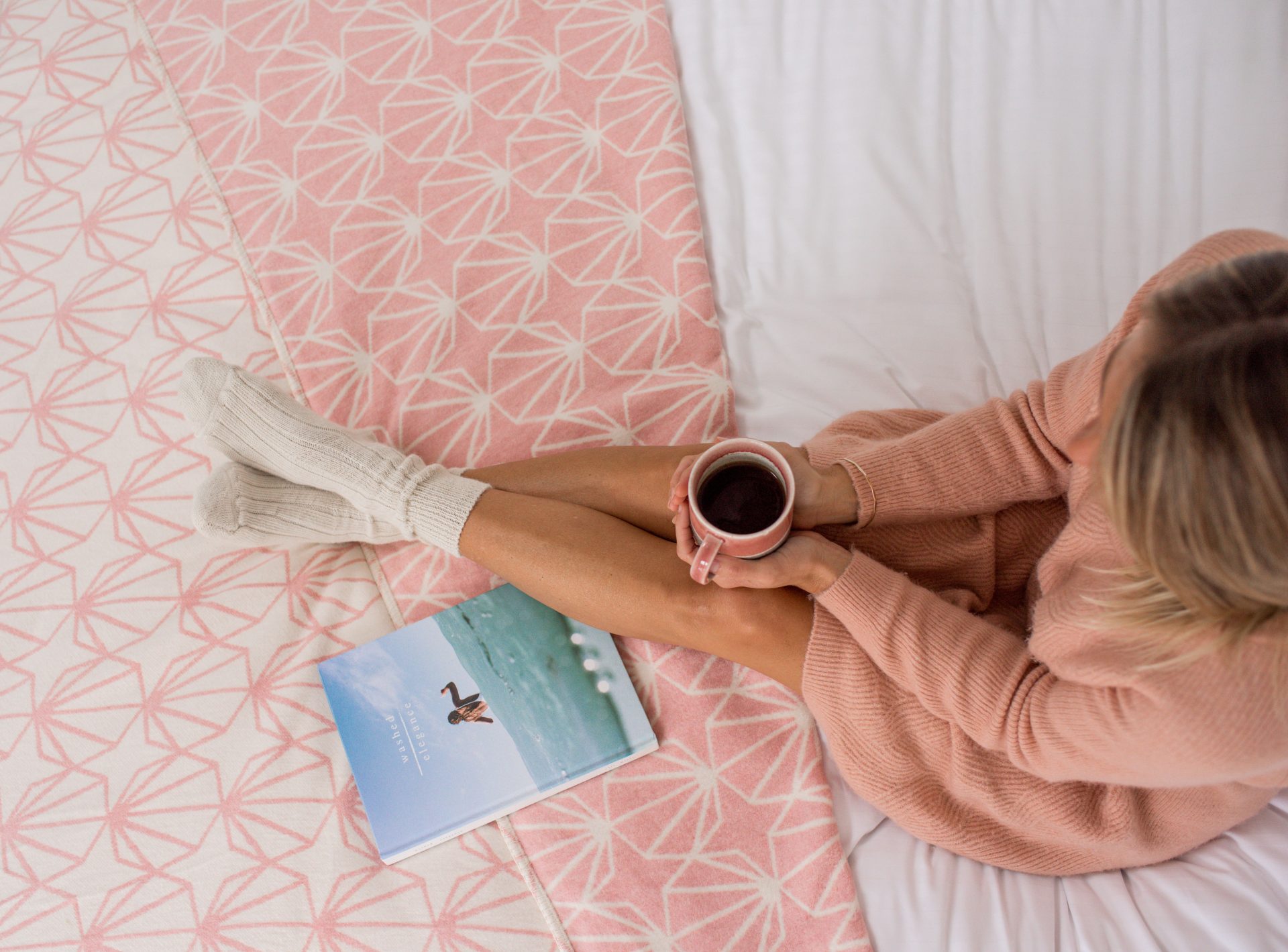 Woman in a pink sweater and white socks sitting on a bed with an Atlantic Blankets pink blanket, holding a mug, next to a book with a bird on the cover.