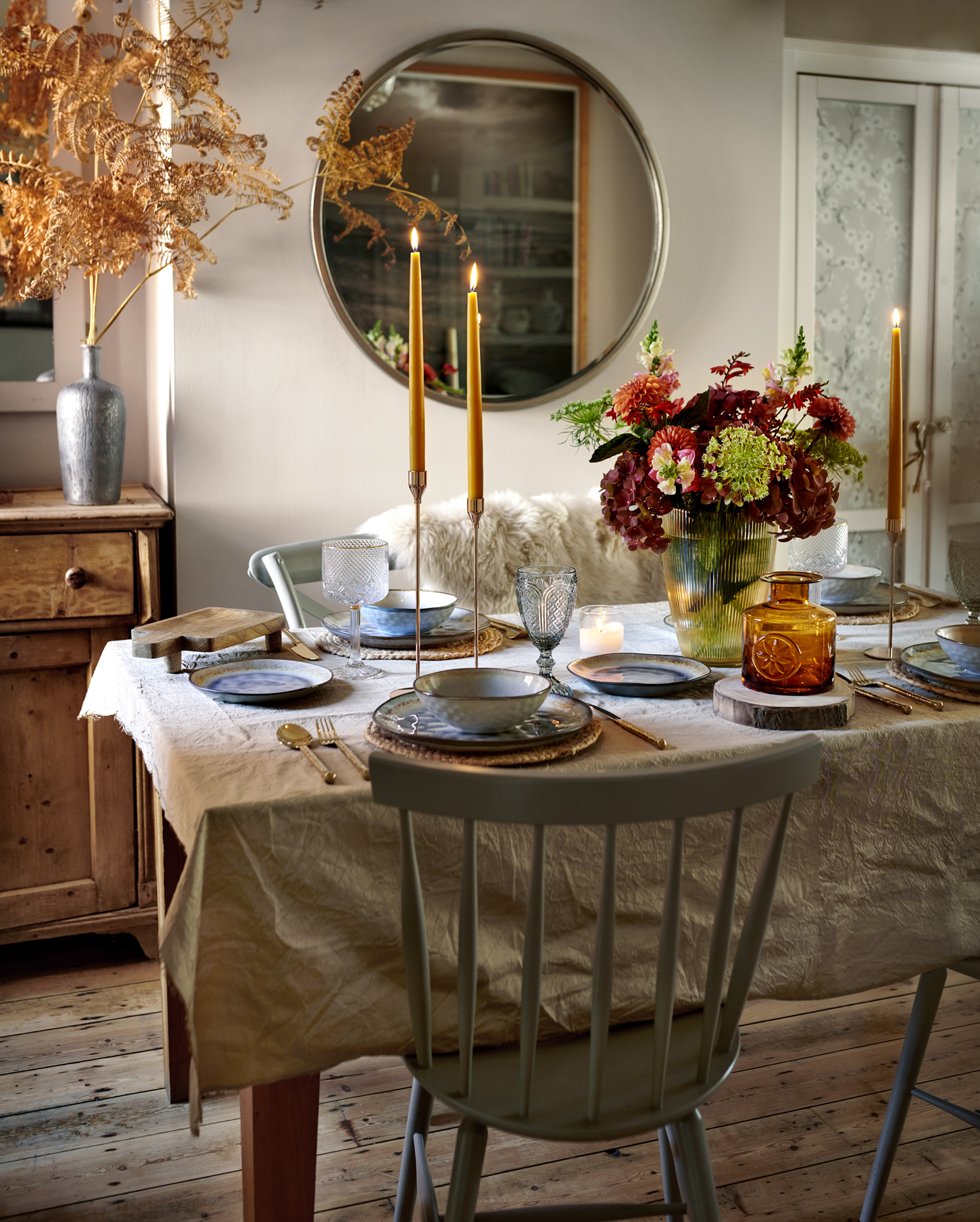 Elegant dining room table set for a meal with candles, ornate silverware, floral arrangements, and reflected in a circular mirror on the wall, creating a cosy home atmosphere.