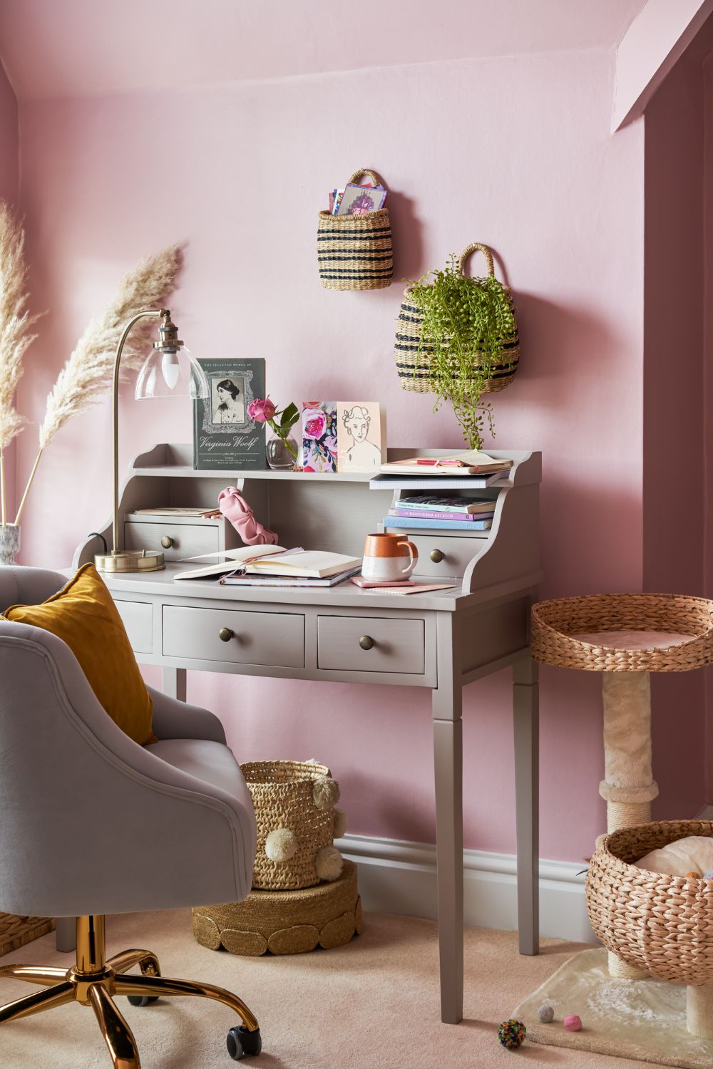 A chic home office with a pink wall, featuring a gray desk with books and decor, a plush chair, and cat accessories including a scratching post.