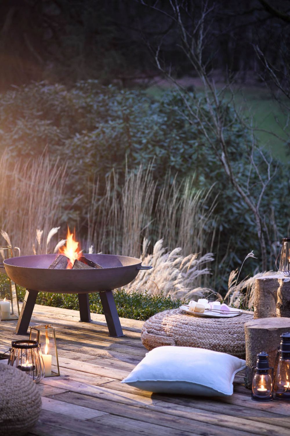 A cozy outdoor living area at dusk with a fire pit, candles, cushions, and wicker stools on a fashionable wooden deck surrounded by tall grass.