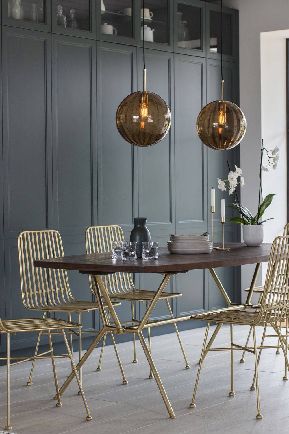 Modern dining area featuring a wooden table, gold-accented chairs, and two hanging glass pendant lights, set against a backdrop of gray cabinetry—perfect for a budget makeover.