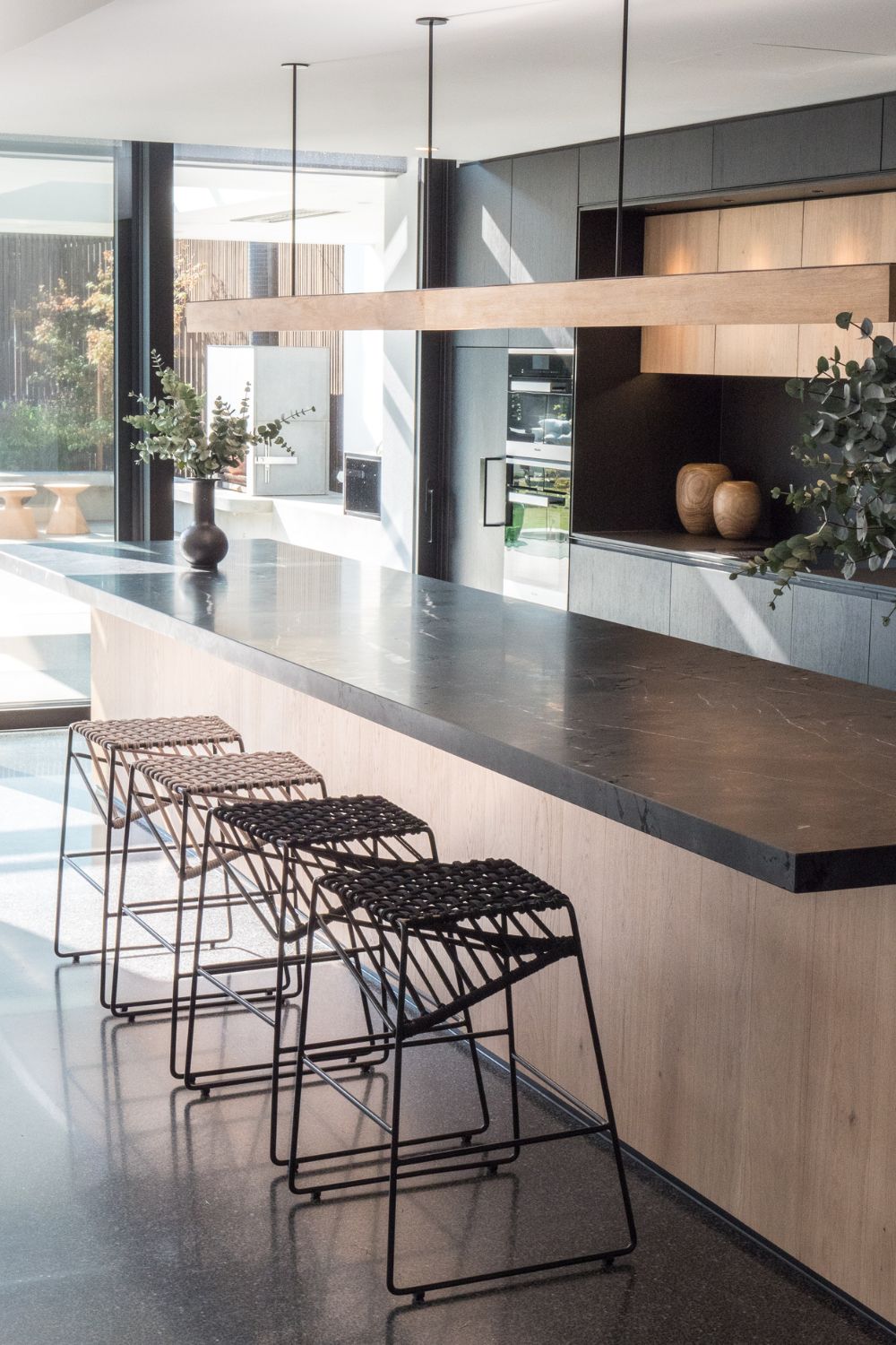 Modern kitchen interior with sleek grey countertops, wooden cabinets, and black bar stools. It showcases an open layout with ample natural light, key factors to consider in design.