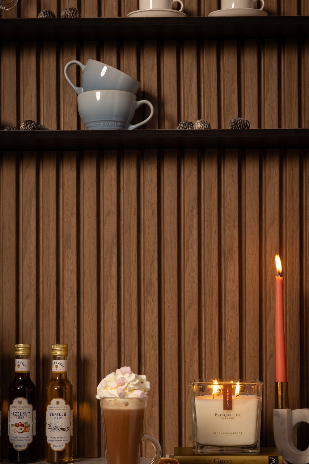 A warm and inviting kitchen scene featuring two stacks of stylish cups on open wooden shelving, a tall glass of hot chocolate topped with whipped cream and marshmallows, flanked by bottles of hazelnut and vanilla syrup. A lit, luxurious Pecksniff's candle provides an ambient glow, with a single lit tall red candle adding a pop of color. The cozy setting is completed with a selection of books, suggesting a relaxed environment for reading and sipping.