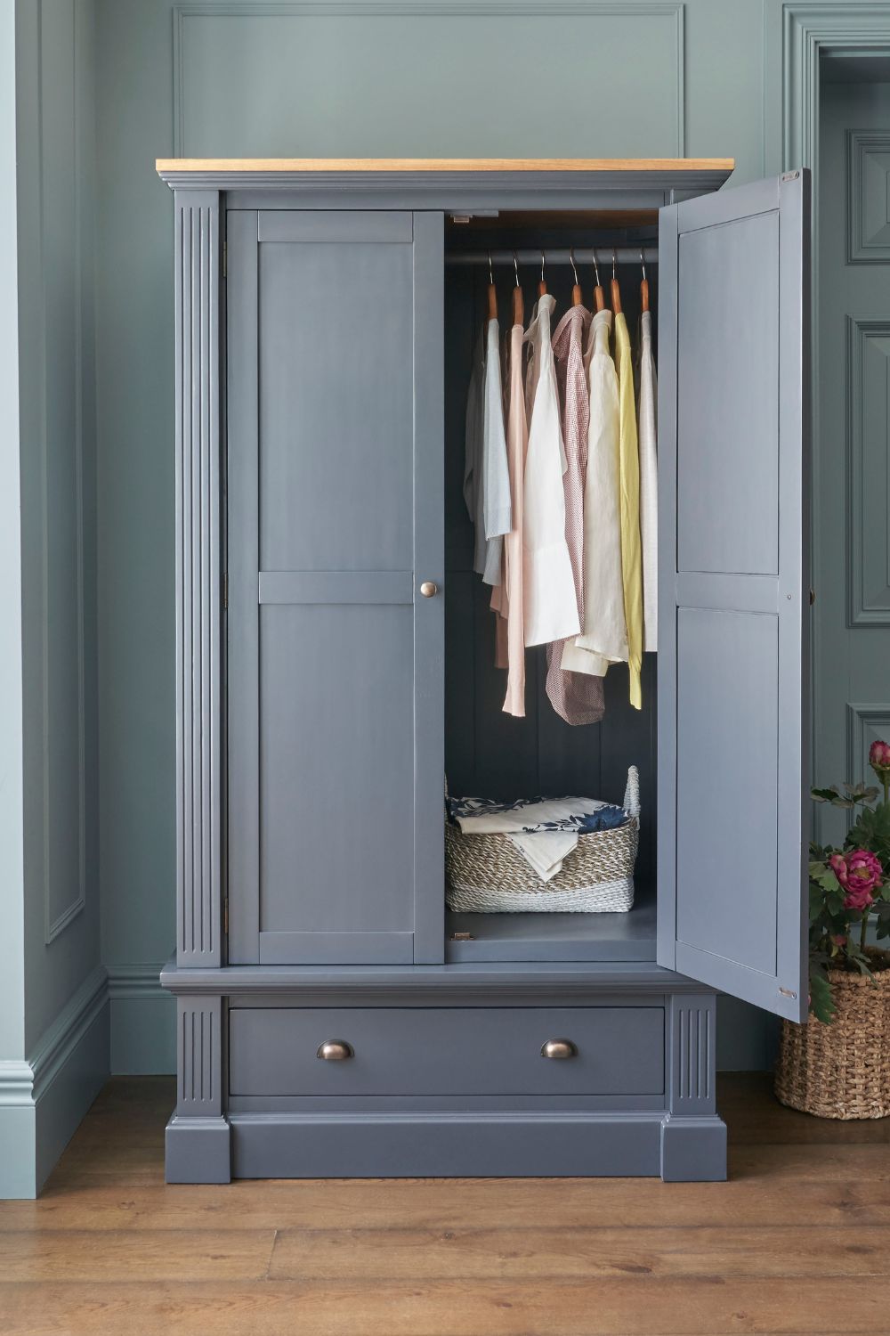 An elegant gray wardrobe with its doors open revealing a neatly organized interior with a selection of hanging pastel-colored clothing and a woven basket on the shelf below. The wardrobe stands against a wall painted in a matching shade of gray, with a natural wood floor and a potted plant adding a touch of greenery to the scene.