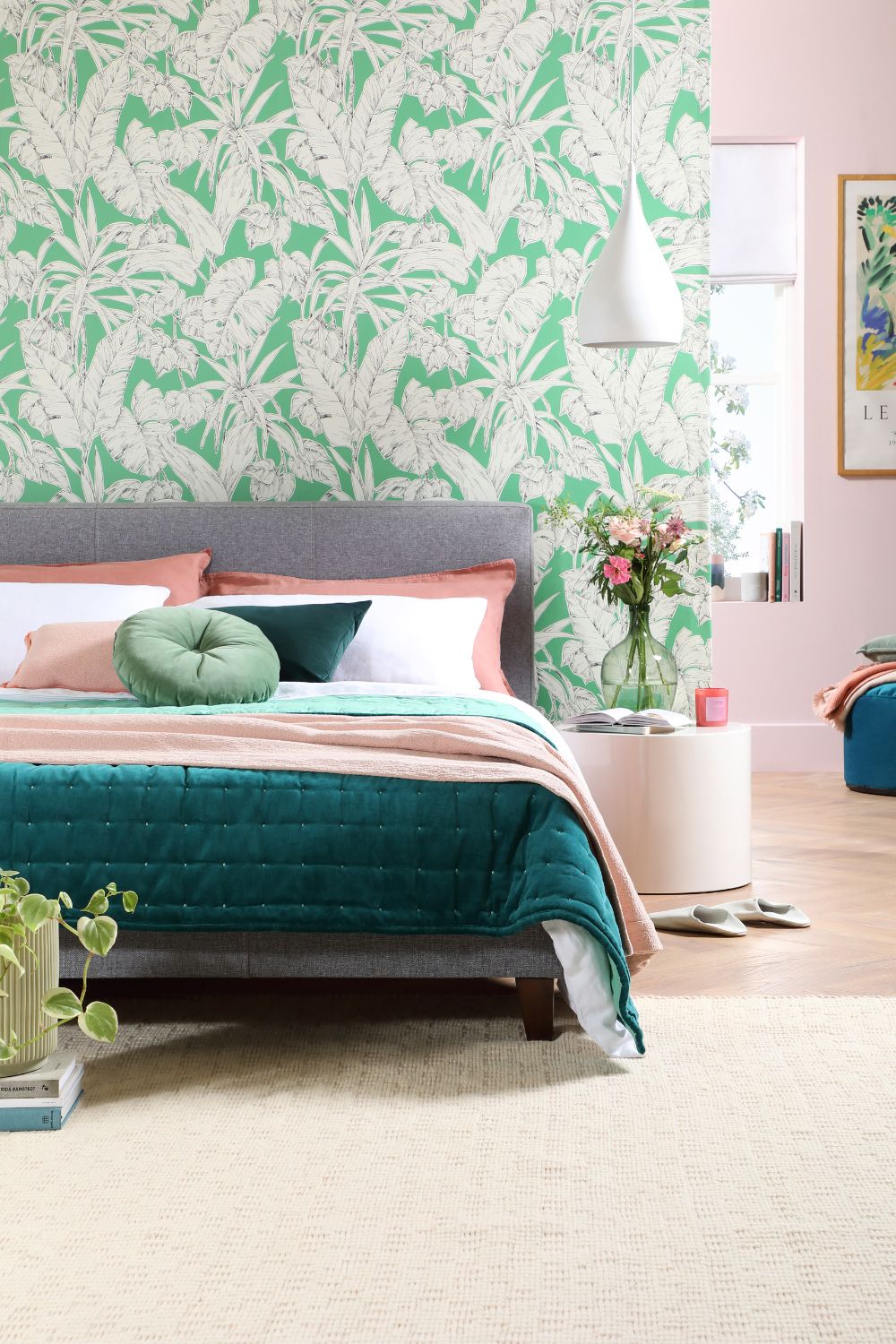 Modern bedroom with a teal bed, pink and green wallpaper, white pendant light, and a beige carpet designed to prevent bed bugs. Decor includes a poster on the wall and a bouquet of flowers on a