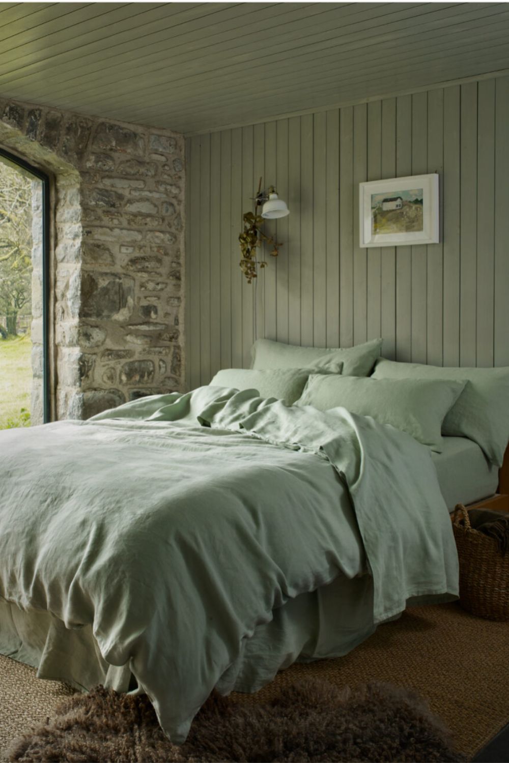 The image shows a bedroom with a prominent sage green accent wall. The wall is composed of vertical panels, providing texture and depth to the space. A stone wall with natural, rough-hewn stones on one side adds a rustic charm. The room is furnished with a bed covered with linens that match the sage green hue of the walls, enhancing the room's monochromatic scheme. A classic white sconce light fixture is mounted on the accent wall, illuminating the room with a soft glow. The ceiling and the remaining walls are painted in soft whites, offering a gentle contrast to the sage green. A framed artwork hangs on the accent wall, adding a personal touch to the space. The overall ambiance is serene and cohesive, with the sage green providing a soothing natural element that is both grounding and refreshing.