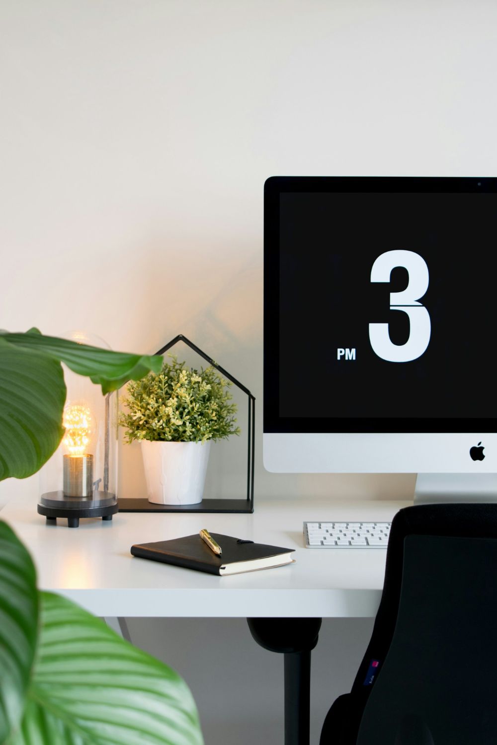 A modern office space with an apple computer displaying the time at 3 pm, accompanied by biophilic design elements and a notebook.