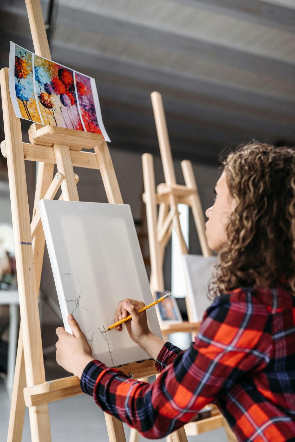 A person with curly hair sketching on a canvas at an easel, with a colorful painting of magical worlds in the background.