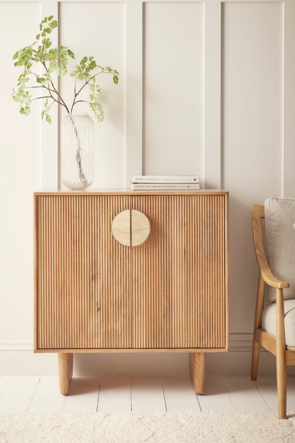 A minimalist wooden cabinet with ribbed doors and circular handles, accompanied by a beige armchair and a vase with greenery on top.