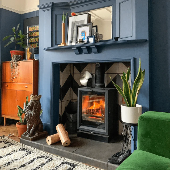 A cozy living room with a lit fireplace using Nestro Heat Logs, surrounded by a blue mantel and decorated with various frames and plants.