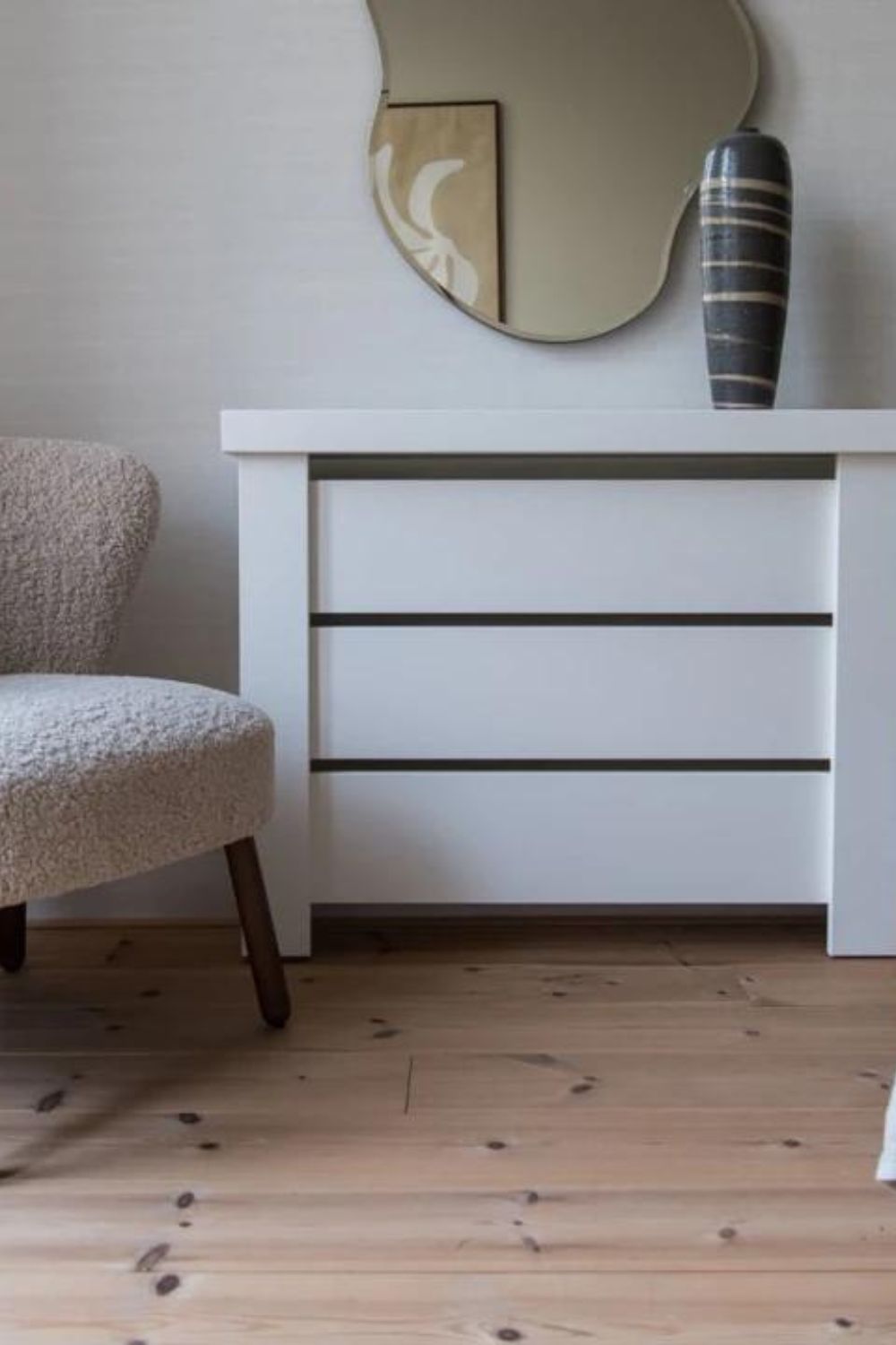 A modern interior featuring a white console table with disguised heating, a beige upholstered chair, a decorative vase, and a round mirror against a textured wall with a light wooden floor.