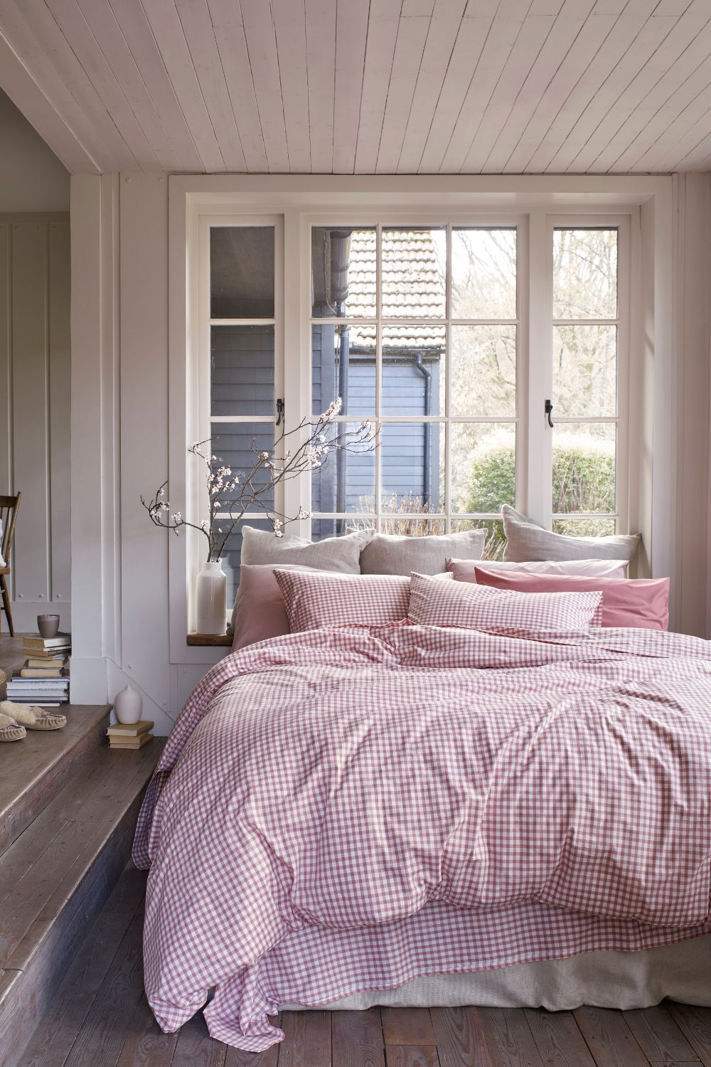 A cozy bedroom with pink checkered bedding, a vase of branches on a window sill, and natural light streaming in through open French windows benefits from small bedroom space hacks that maximize comfort and style.