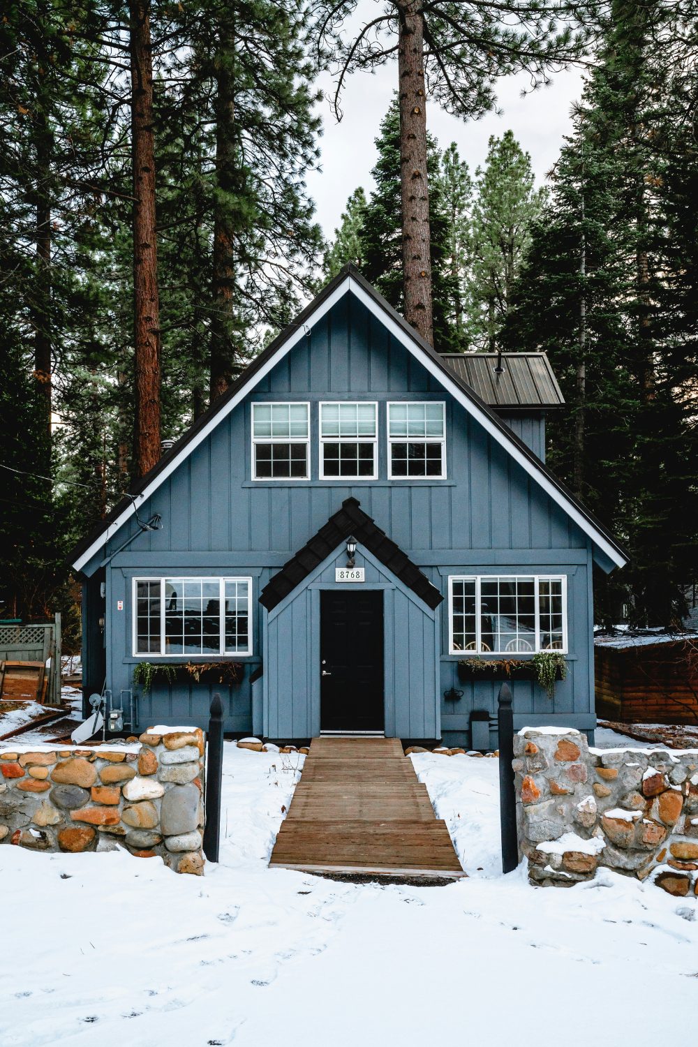 A blue wooden house in a snowy wood bordered by stone walls