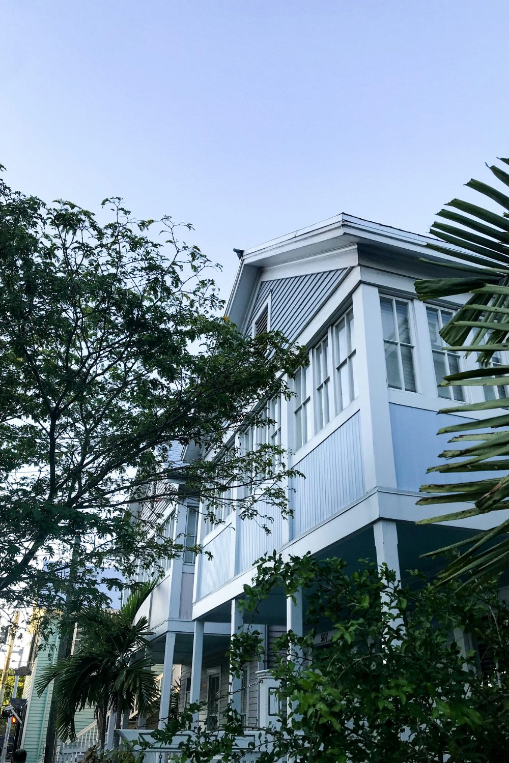 Blue and white wooden house with a first floor raised on stilts flanked by foliage