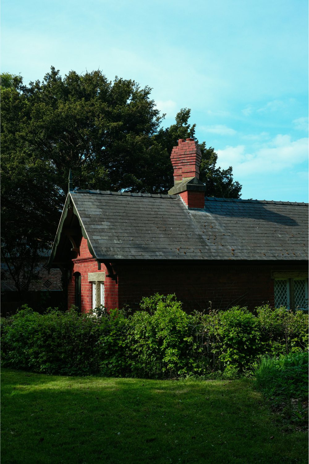 Red brick house with black roof surrounded by hedge under a bright blue sky