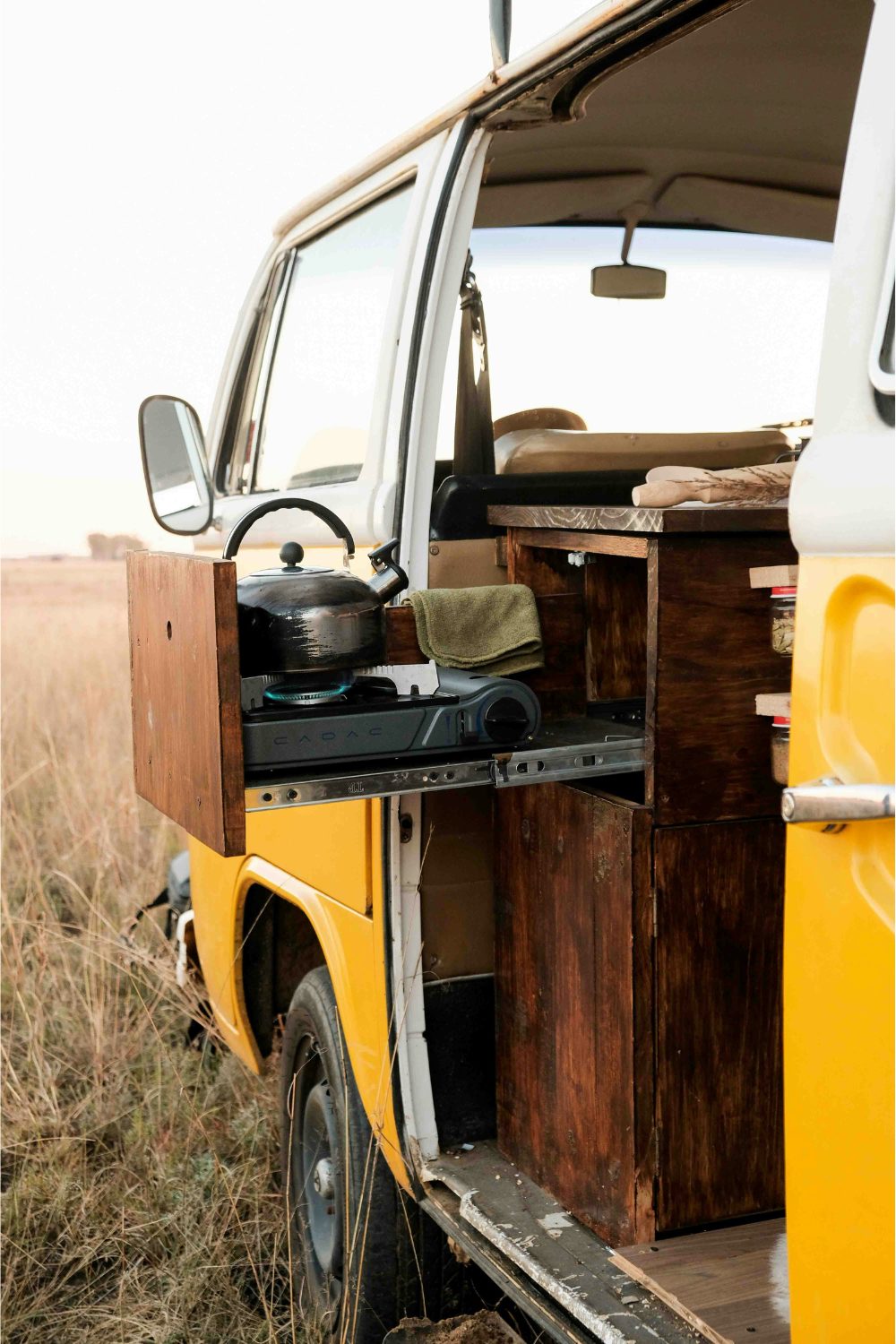 Side of a yellow campervan in a grassy field with a wooden pullout drawer containing a burner and kettle